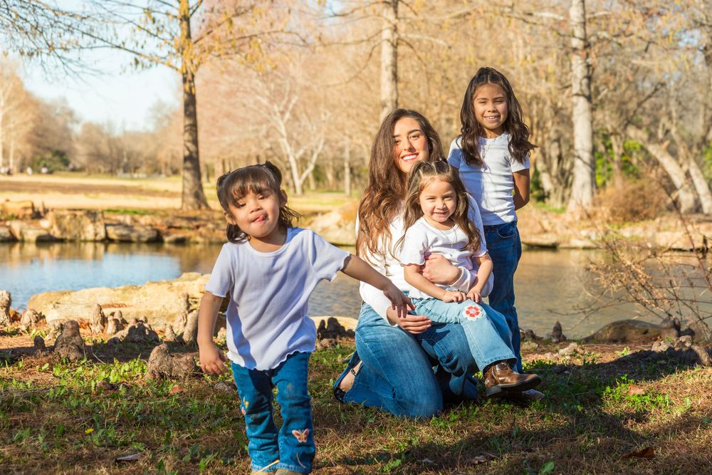 A mother poses outdoors with her three young children near a pond in a sunny park setting.