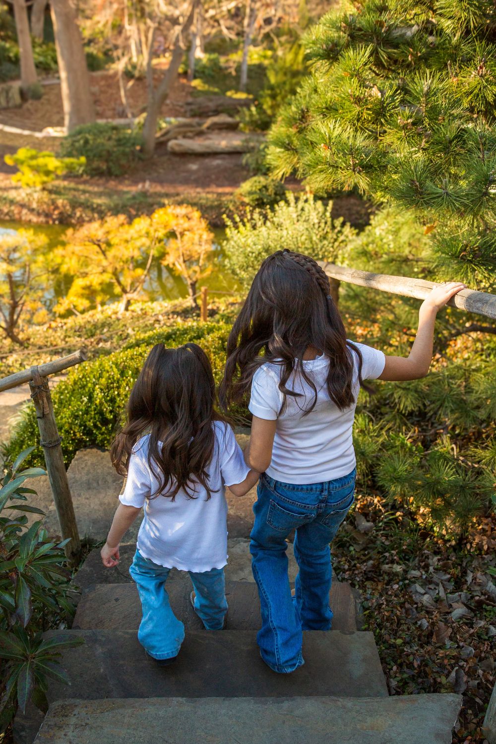 Two young girls with long dark hair in white shirts and jeans walk down garden steps surrounded by lush greenery.
