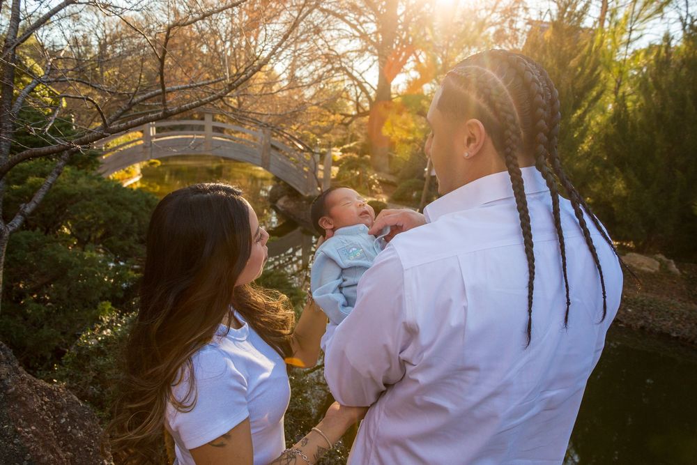 A couple admires their newborn baby in a sunlit Japanese garden with a wooden bridge and autumn trees.