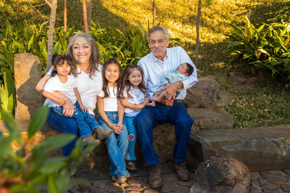 Grandparents sit outdoors with four grandchildren, including a newborn, in a green garden.