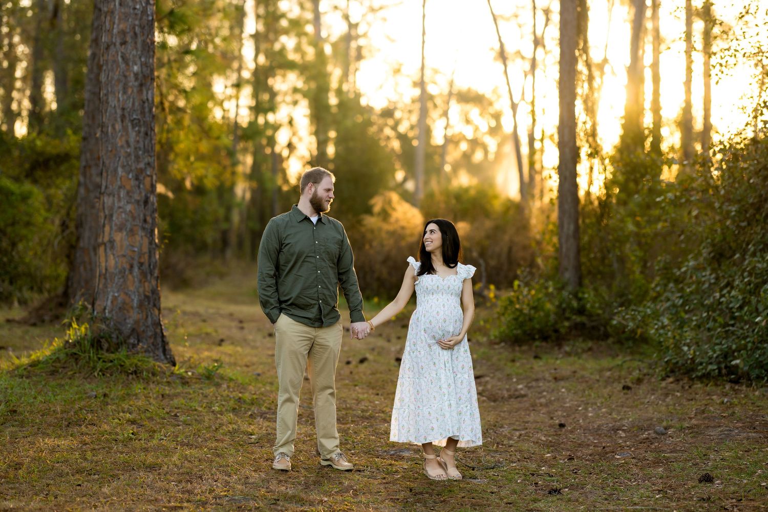Pregnant couple holding hands in a sunlit forest clearing, woman in white lace dress, man in green shirt.
