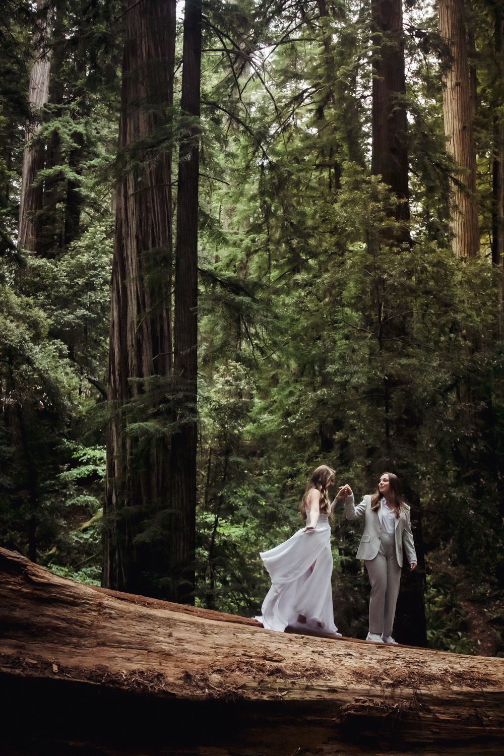 Wedding couple standing among towering redwood trees in a lush forest, bride in white gown and her bride in suit.