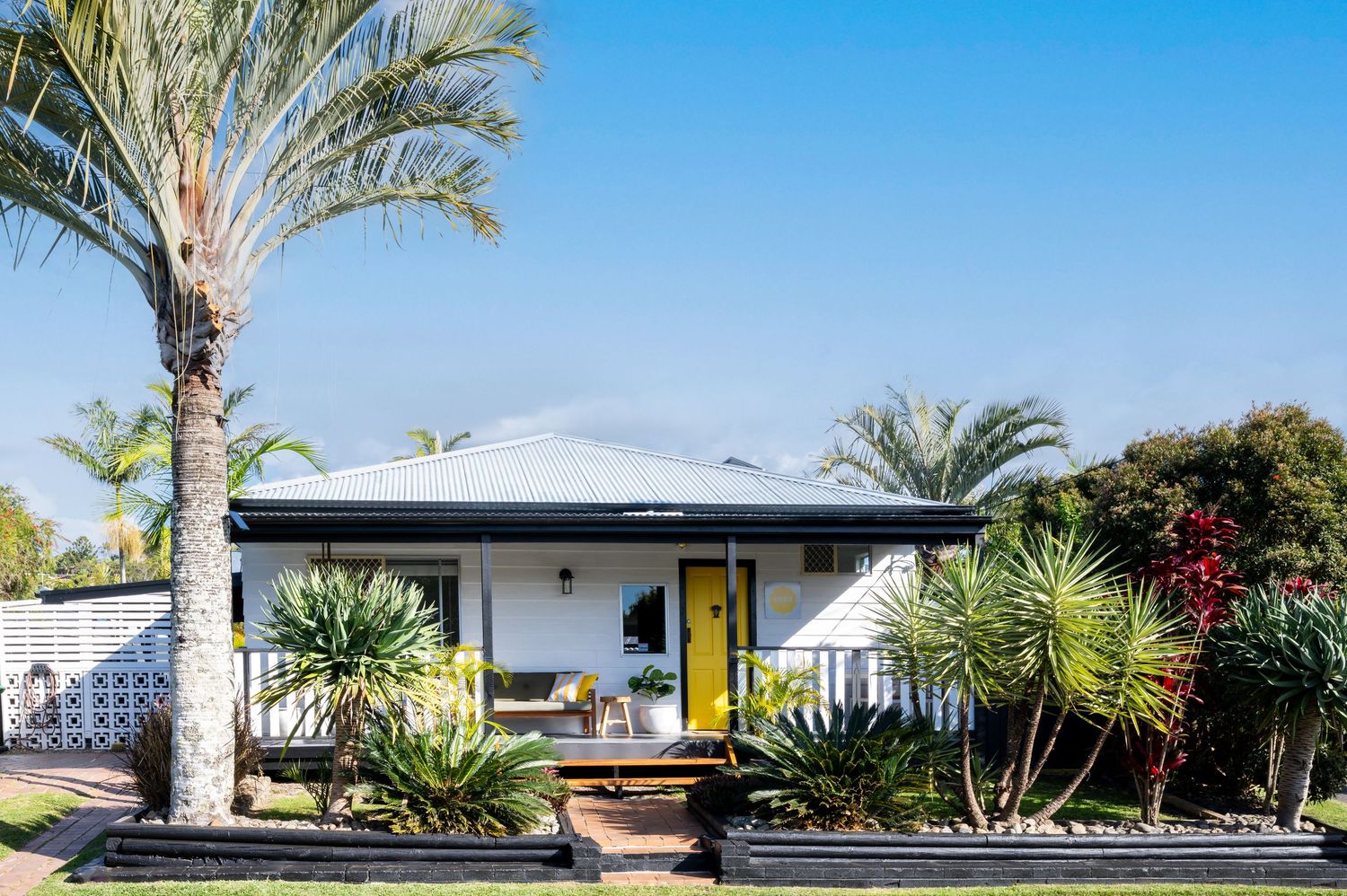 White bungalow with yellow front door, tropical palms, and lush garden landscaping under a clear blue sky.