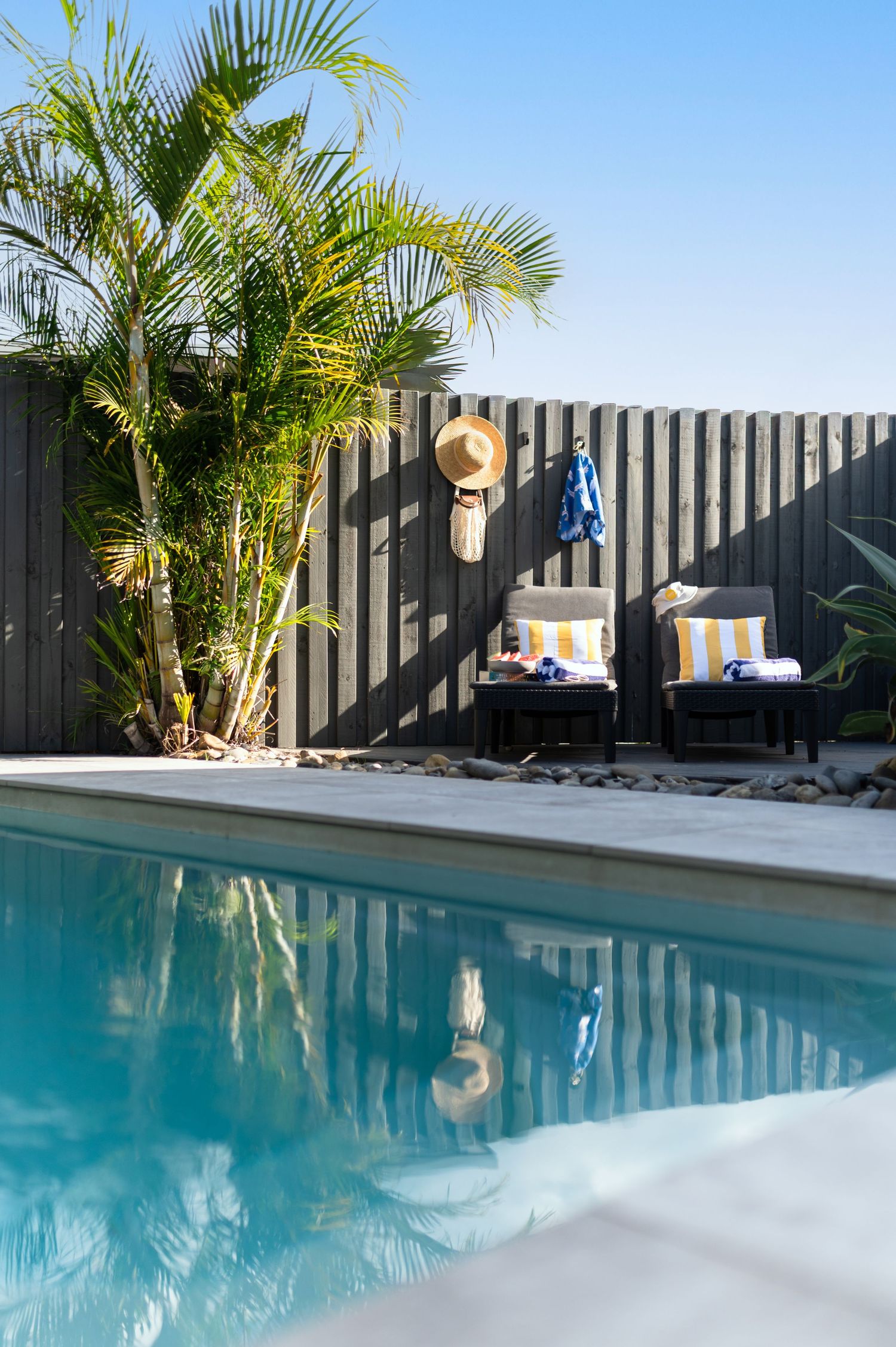 Sunny backyard pool with lounge chairs, striped cushions, a straw hat on a dark wood fence, and lush palm trees.