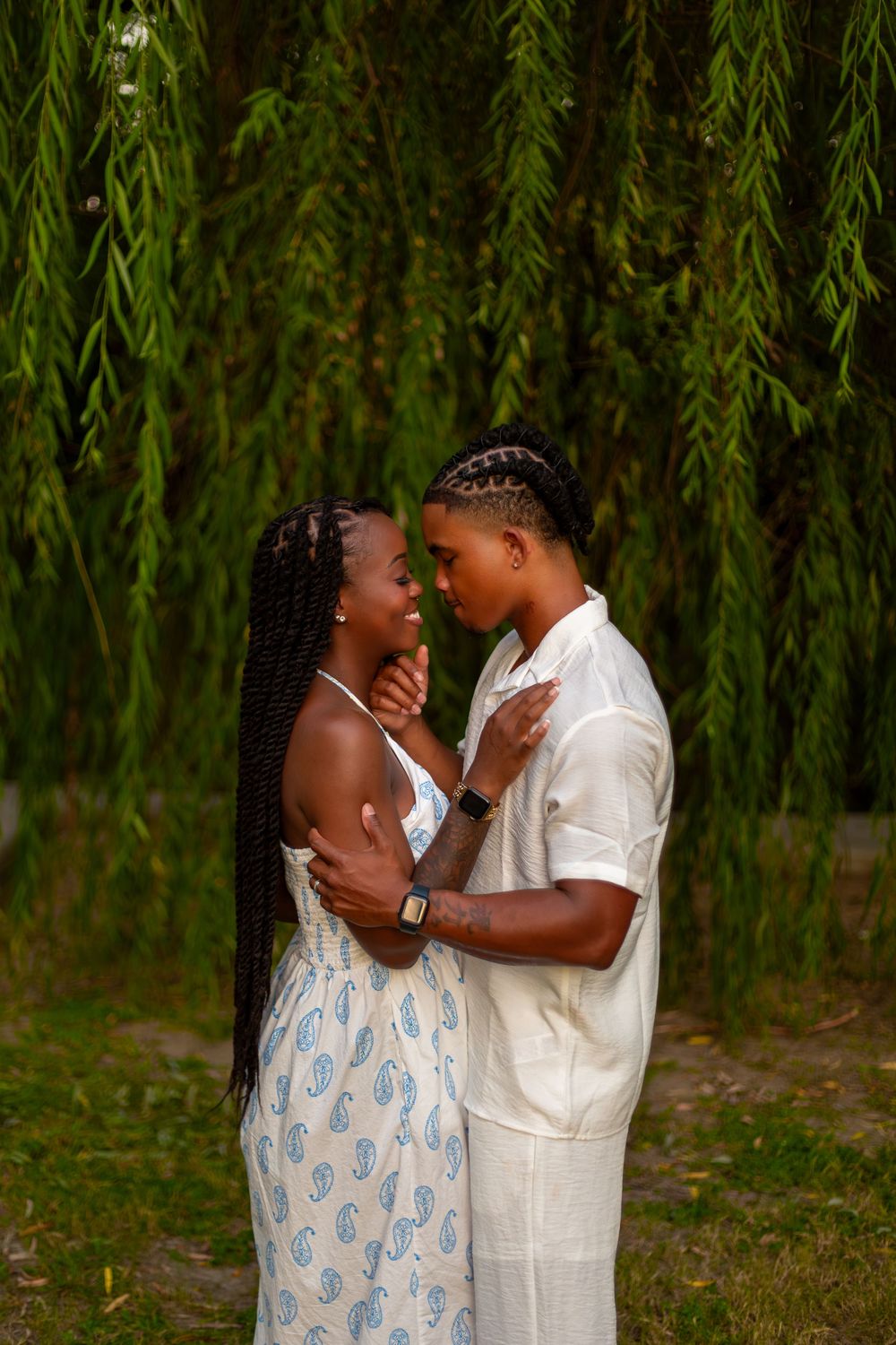 A Black couple shares a romantic moment under a weeping willow tree, dressed in elegant white and blue outfits.