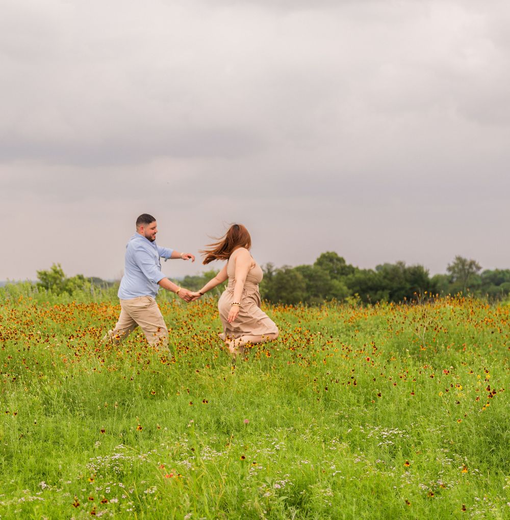Couple dancing joyfully in a wildflower meadow, holding hands with flowing dress and cloudy sky backdrop.