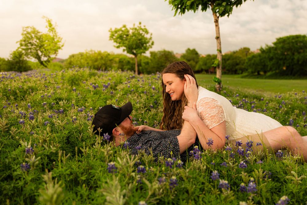 Couple lying in a field of bluebonnets at golden hour, sharing a romantic moment among wildflowers and trees.