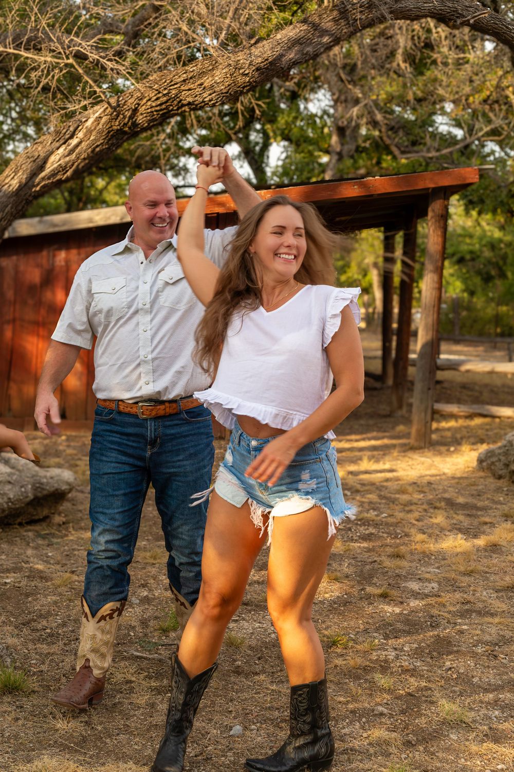 A smiling couple in cowboy boots dance outdoors near a red barn, woman in denim shorts and white top, man in jeans.