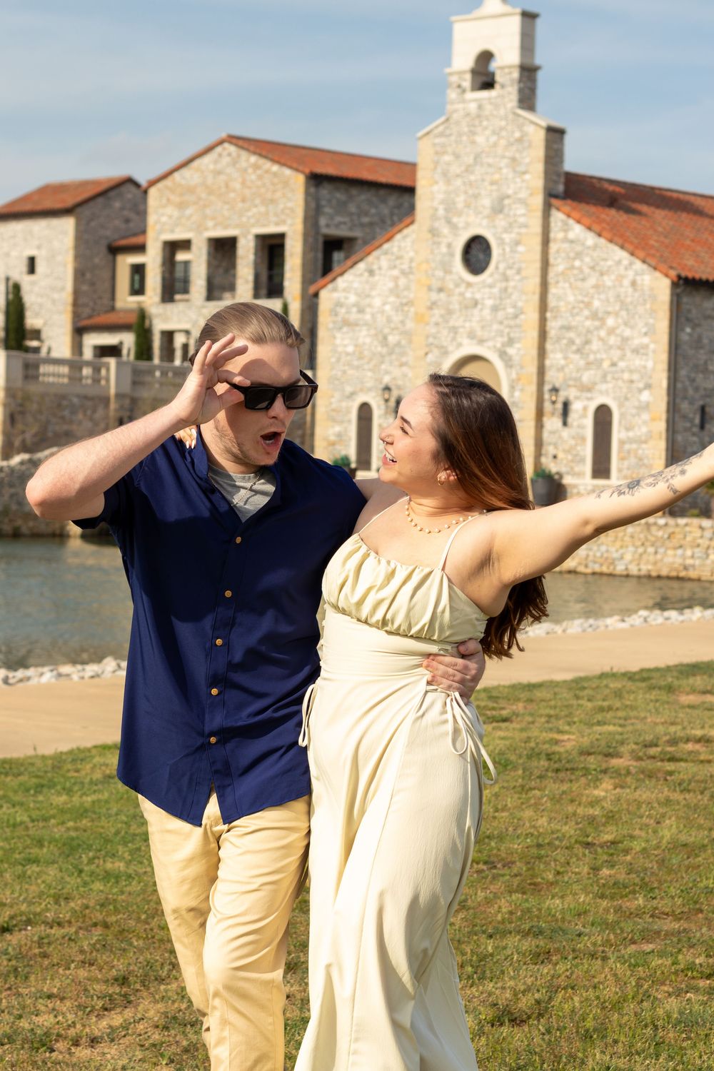 A joyful couple walks outdoors near a stone church, woman in cream dress, man in navy shirt wearing sunglasses.