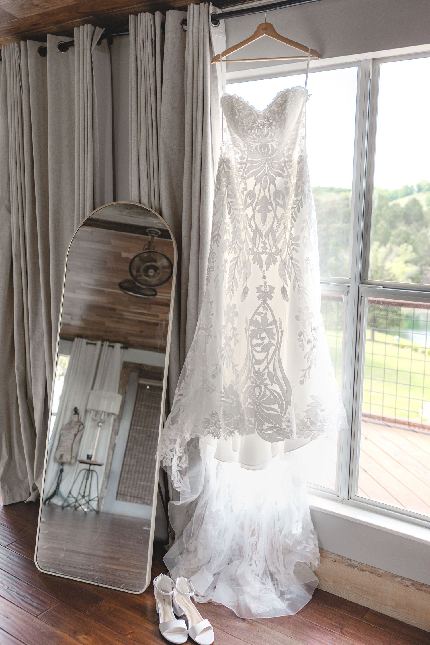 Elegant lace wedding dress hanging by a window with a full-length mirror and bridal heels on a hardwood floor.