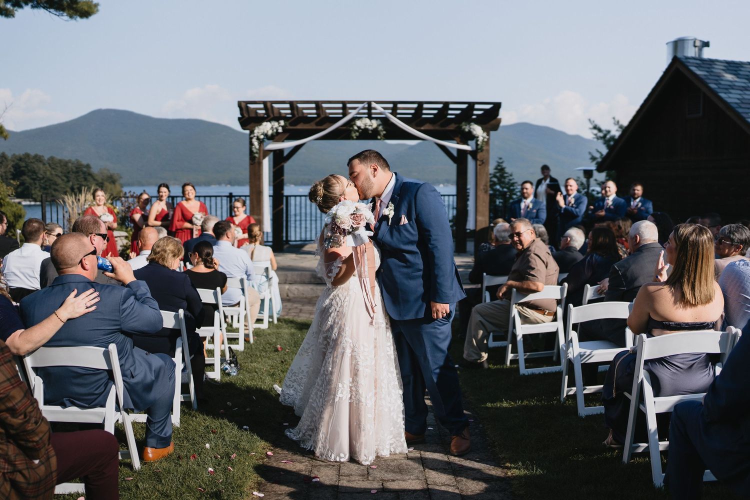 Outdoor wedding ceremony by a lakeside with mountain views, floral barrel aisle markers, and guests seated in white chairs.