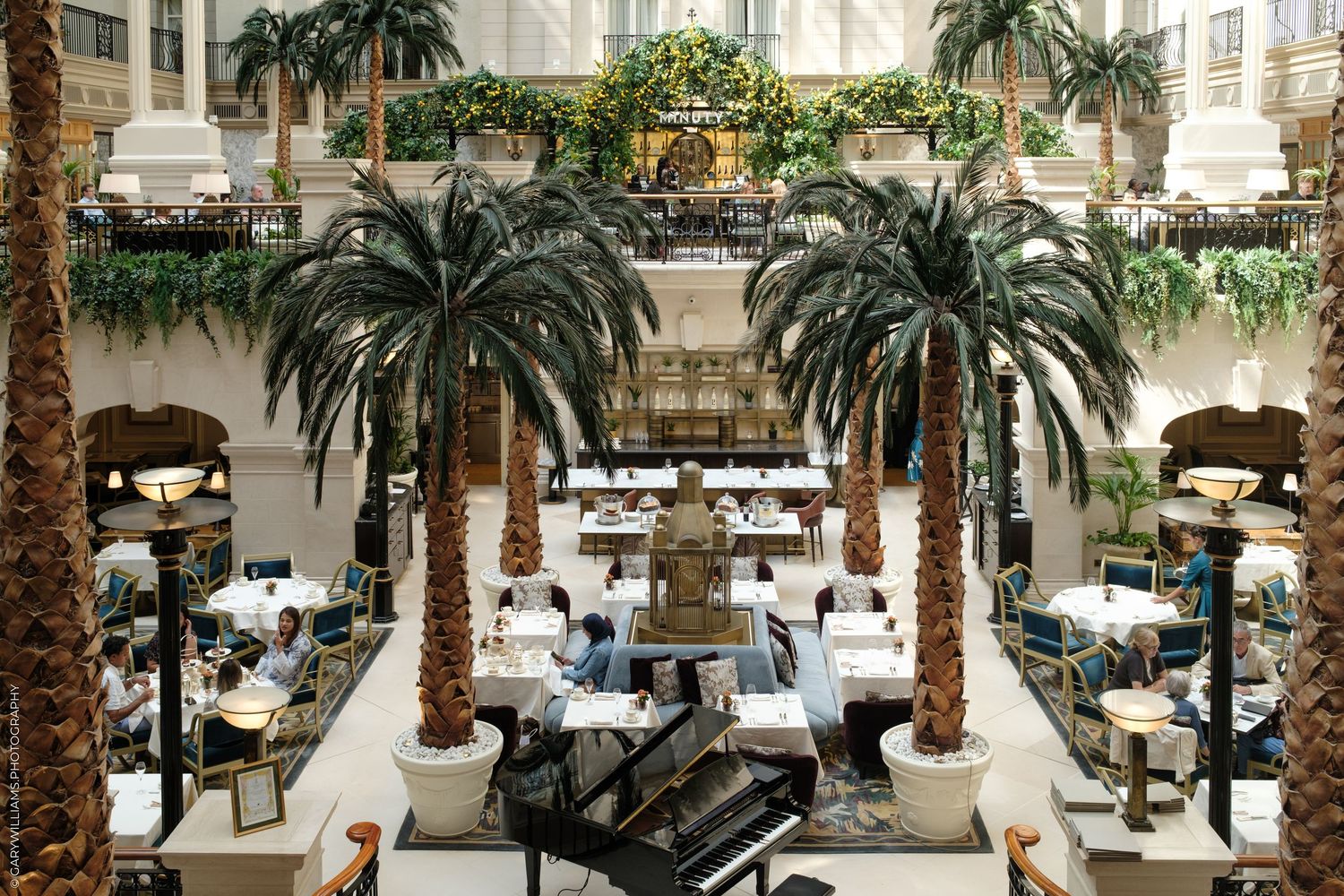 Elegant hotel atrium with tall palm trees, a grand piano, lush greenery, and guests dining in a luxurious multi-level space.