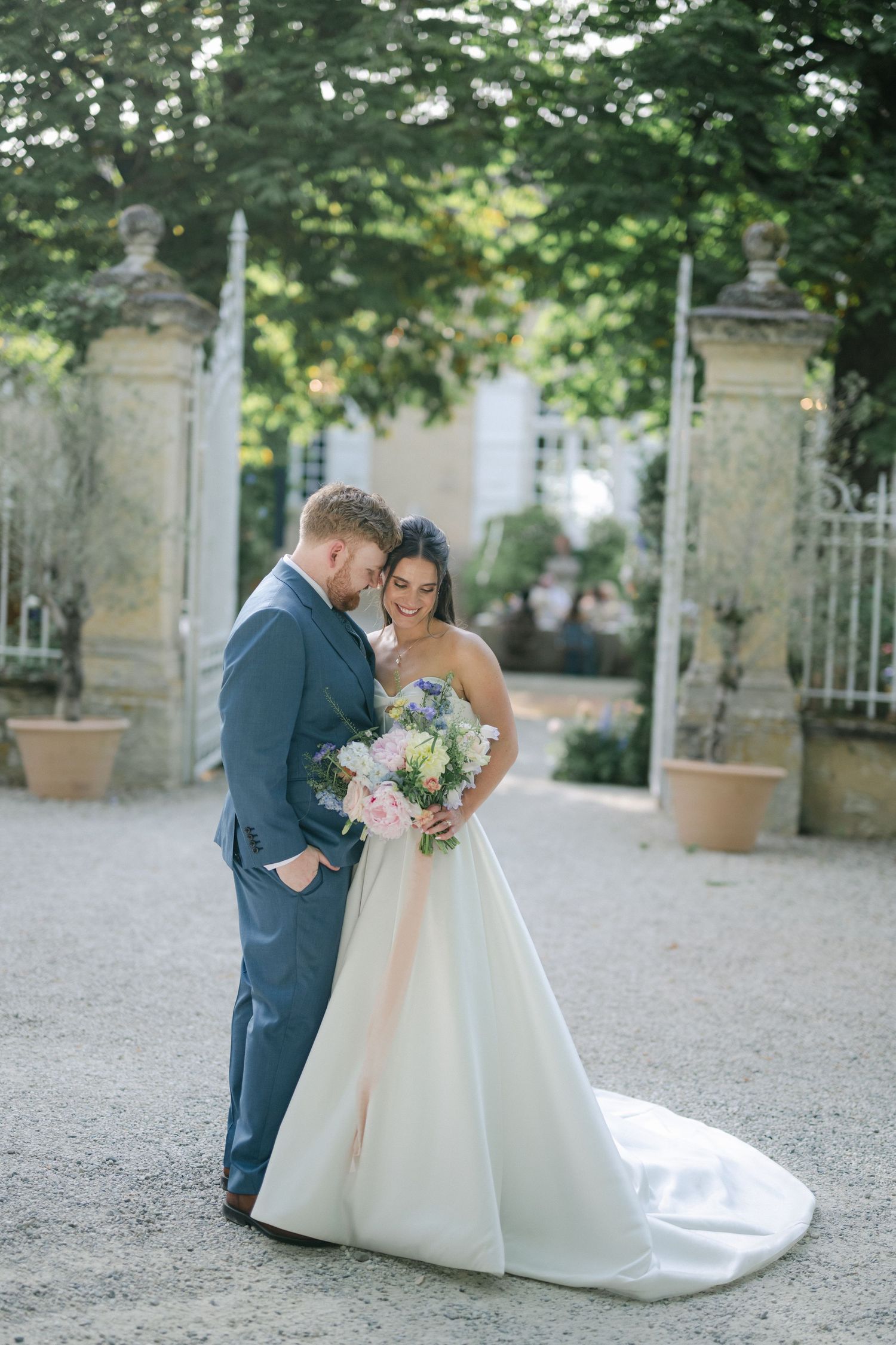 A smiling bride in a white strapless gown holds hands with a groom in a blue suit outdoors at a wedding venue.