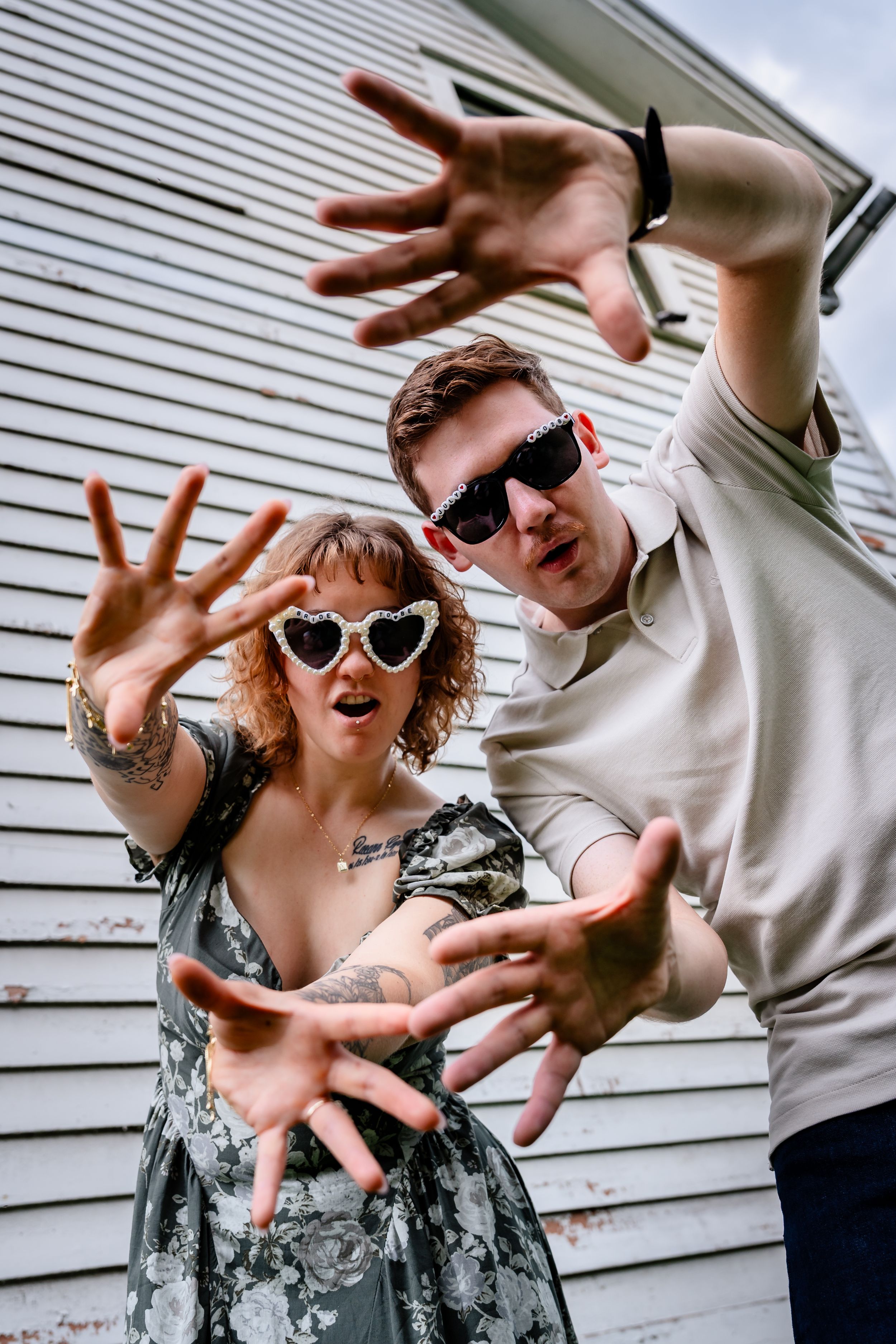 Two people in sunglasses reaching toward camera with clawed hands, posing dramatically outdoors near a white building.