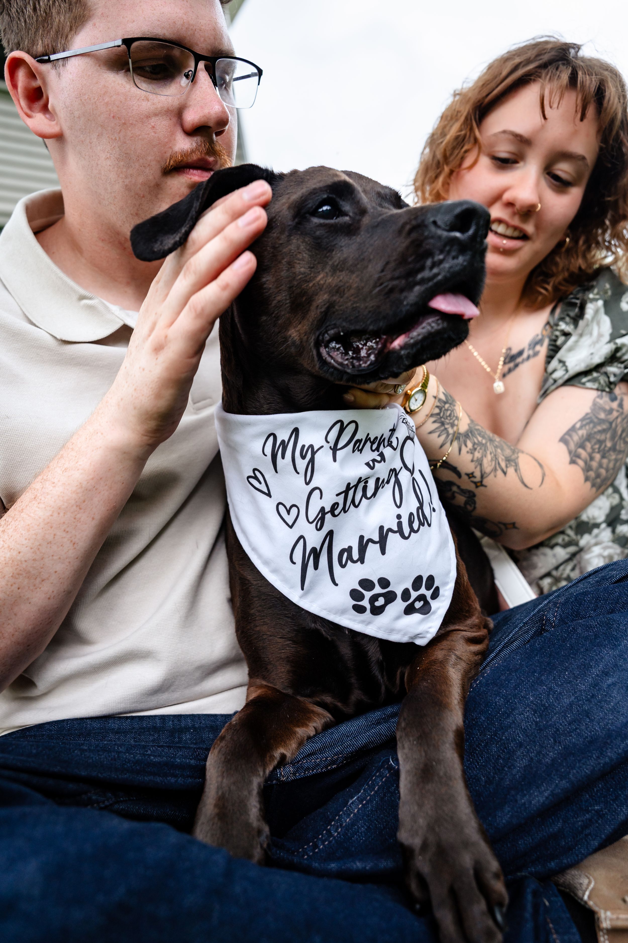 Couple petting their brown dog wearing a white bandana that reads My Parents Are Getting Married during engagement photos.