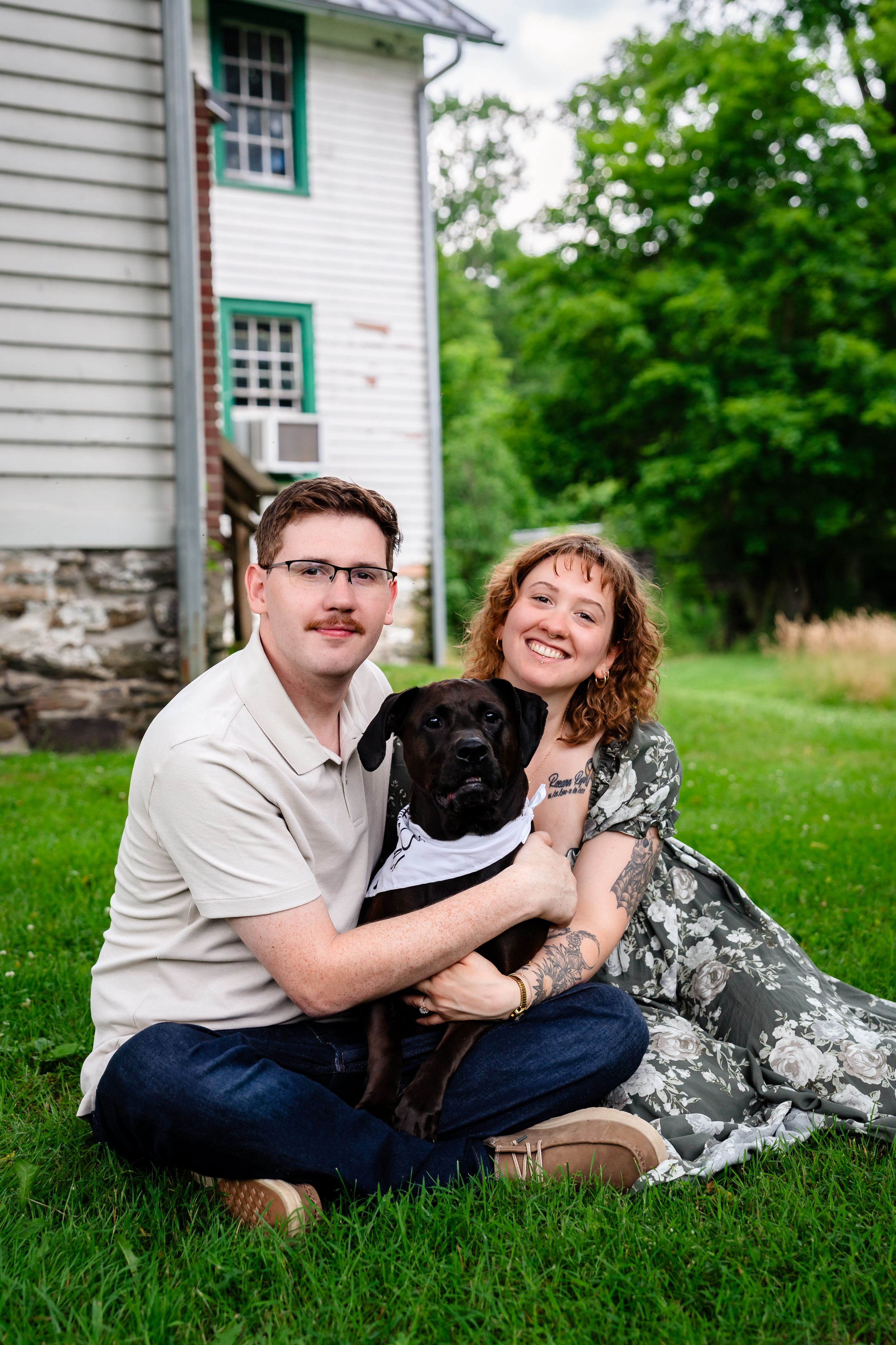 Smiling couple sitting on grass with their black Labrador wearing a bandana outside a white house.