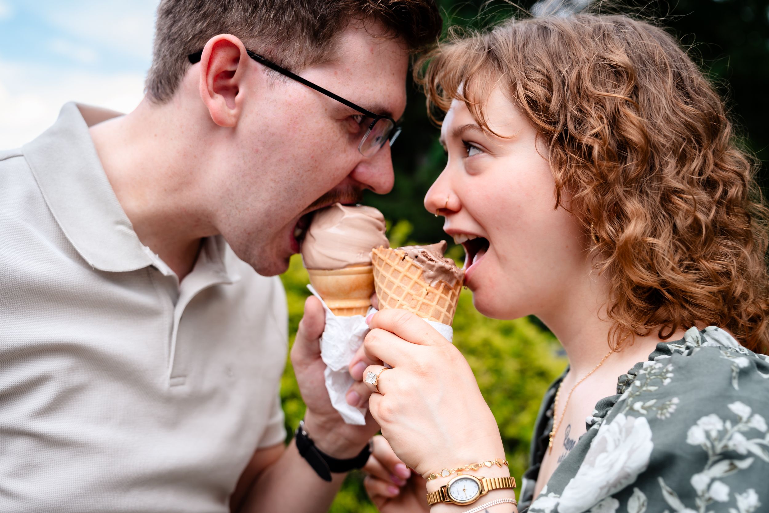 A playful couple laughs while eating ice cream cones together outdoors on a sunny day.
