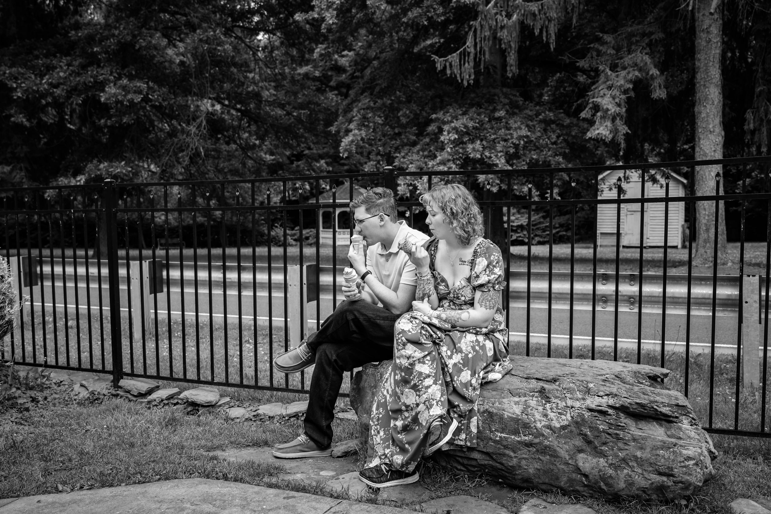 Black and white photo of a woman and young boy sitting together on rocks near an iron fence outdoors.