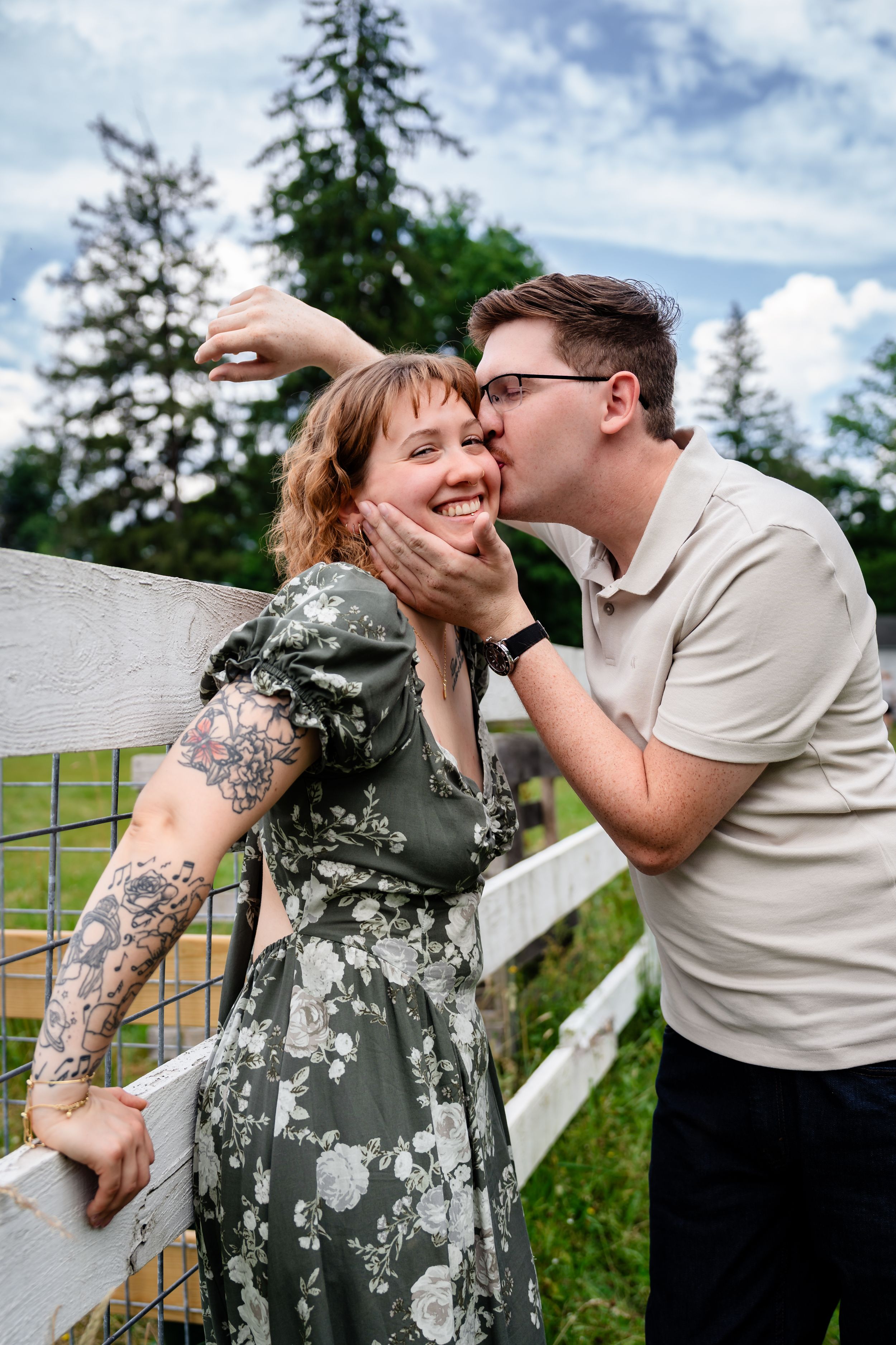 A couple shares a romantic kiss by a white fence, the woman in a floral dress with tattoos smiling joyfully outdoors.