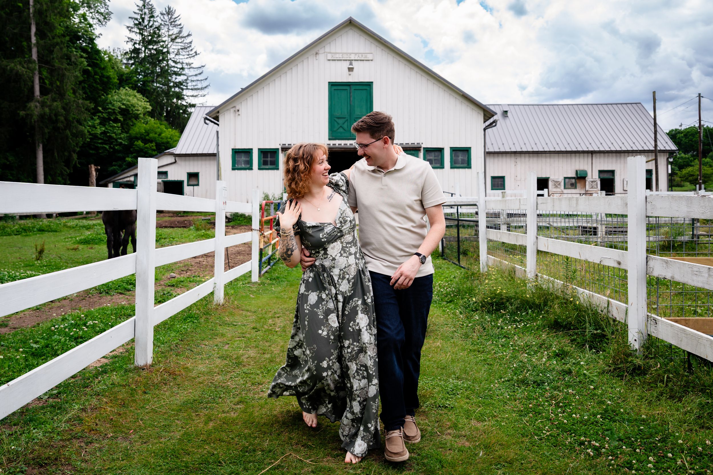 Couple walking hand in hand by white fence at a farm with a white barn in the background.