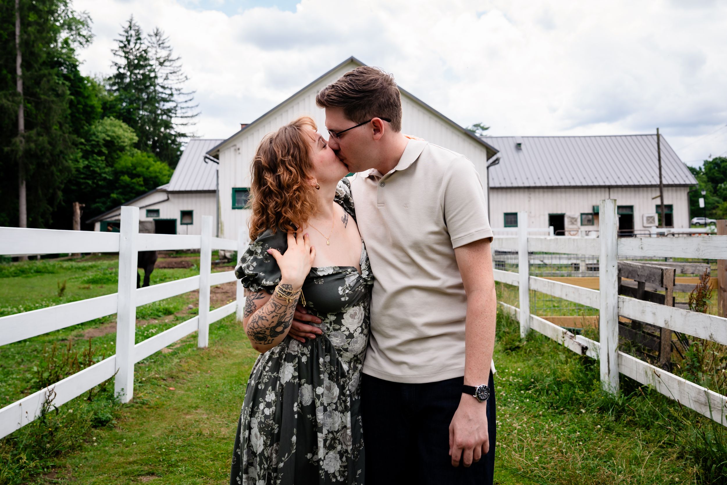 Couple sharing a kiss near white fence at a farm with a barn in the background on a cloudy day.