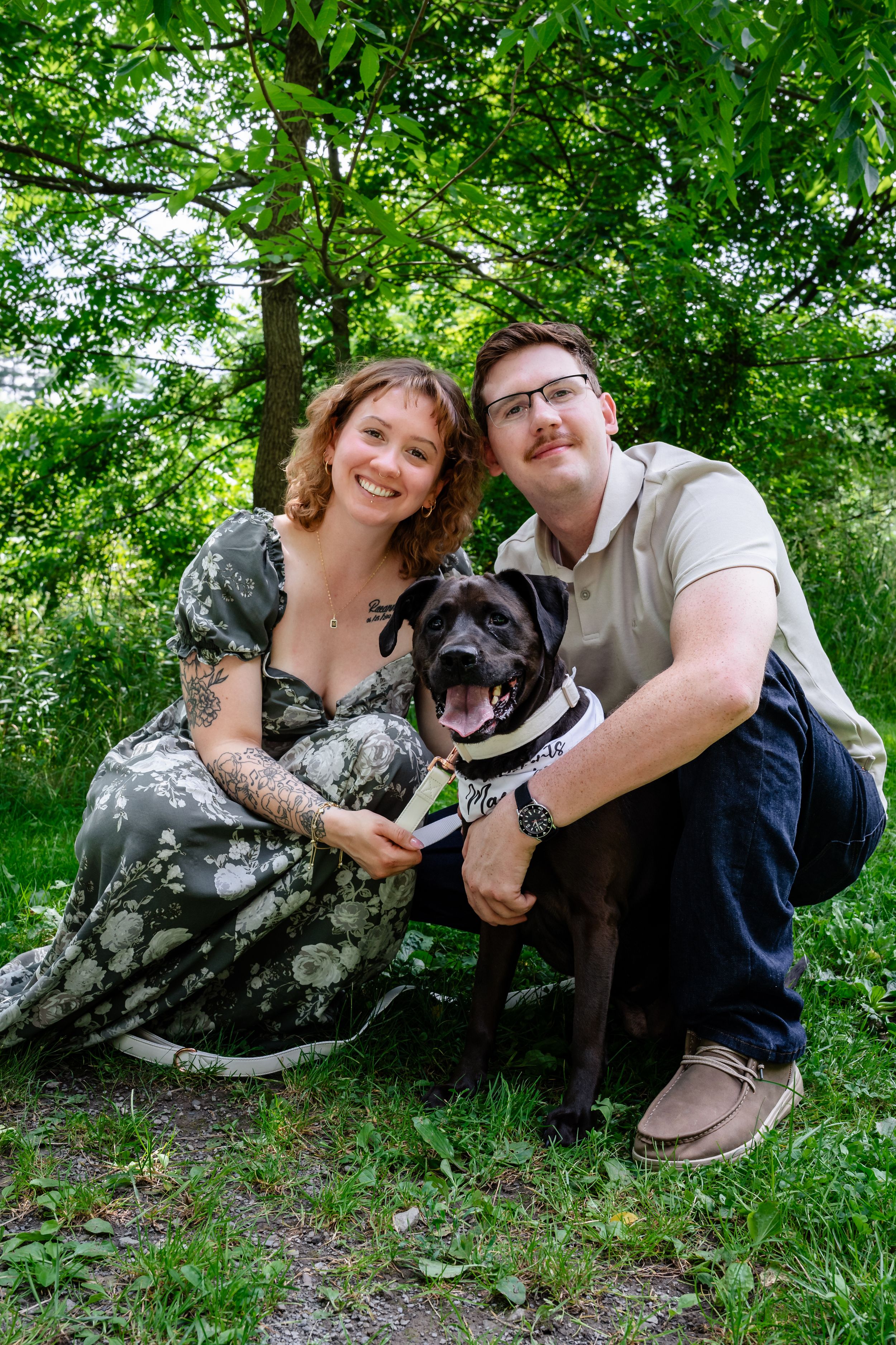 A smiling couple poses outdoors with a black Cane Corso dog, surrounded by lush green trees.