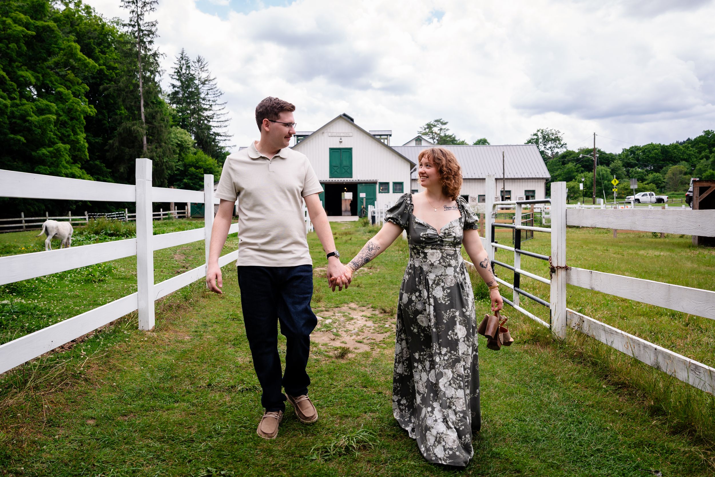 Couple holding hands walking near white fence at farm with barn in background on cloudy day.