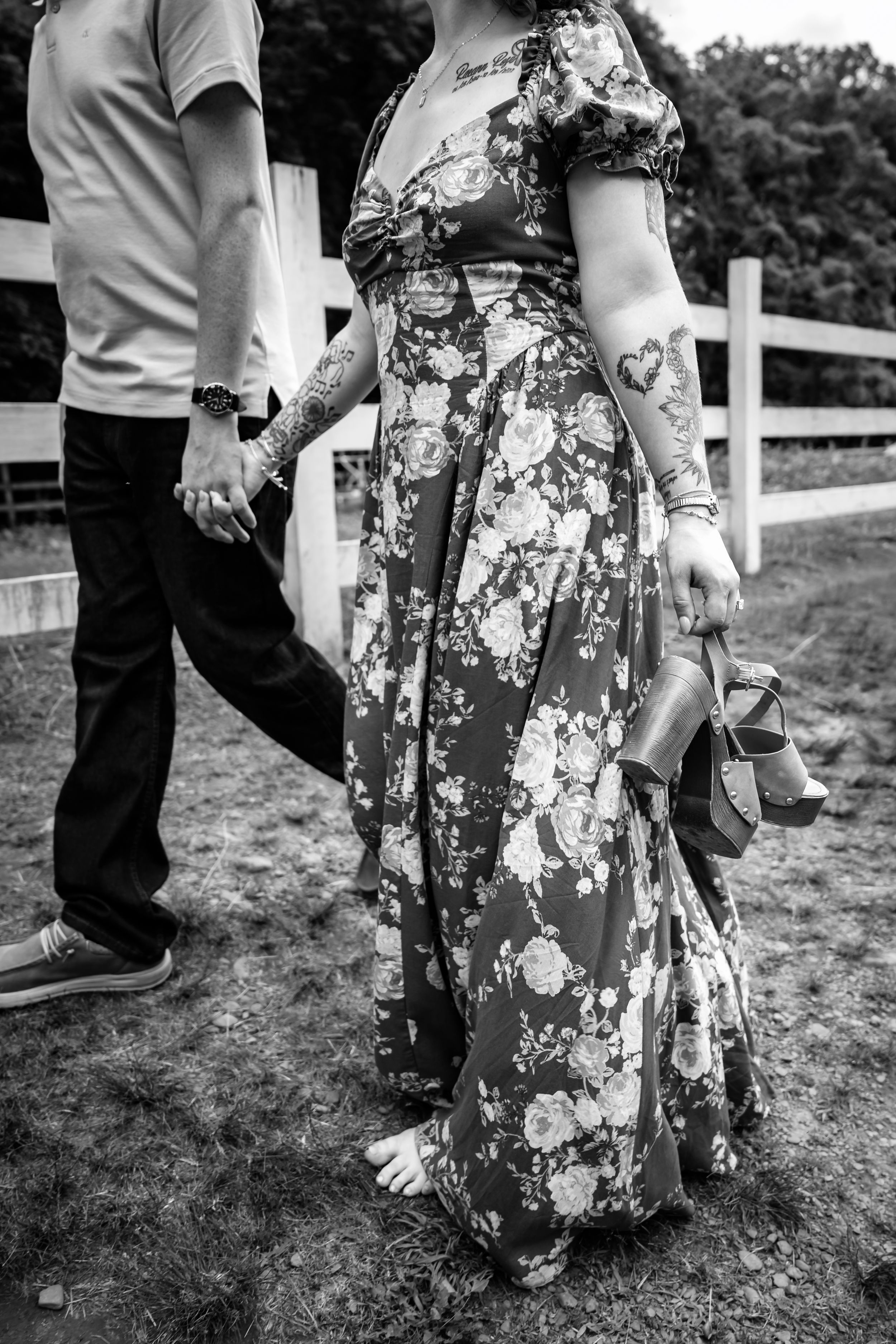 Black and white photo of a tattooed woman in a floral dress holding shoes, walking hand in hand with a man by a white fence.