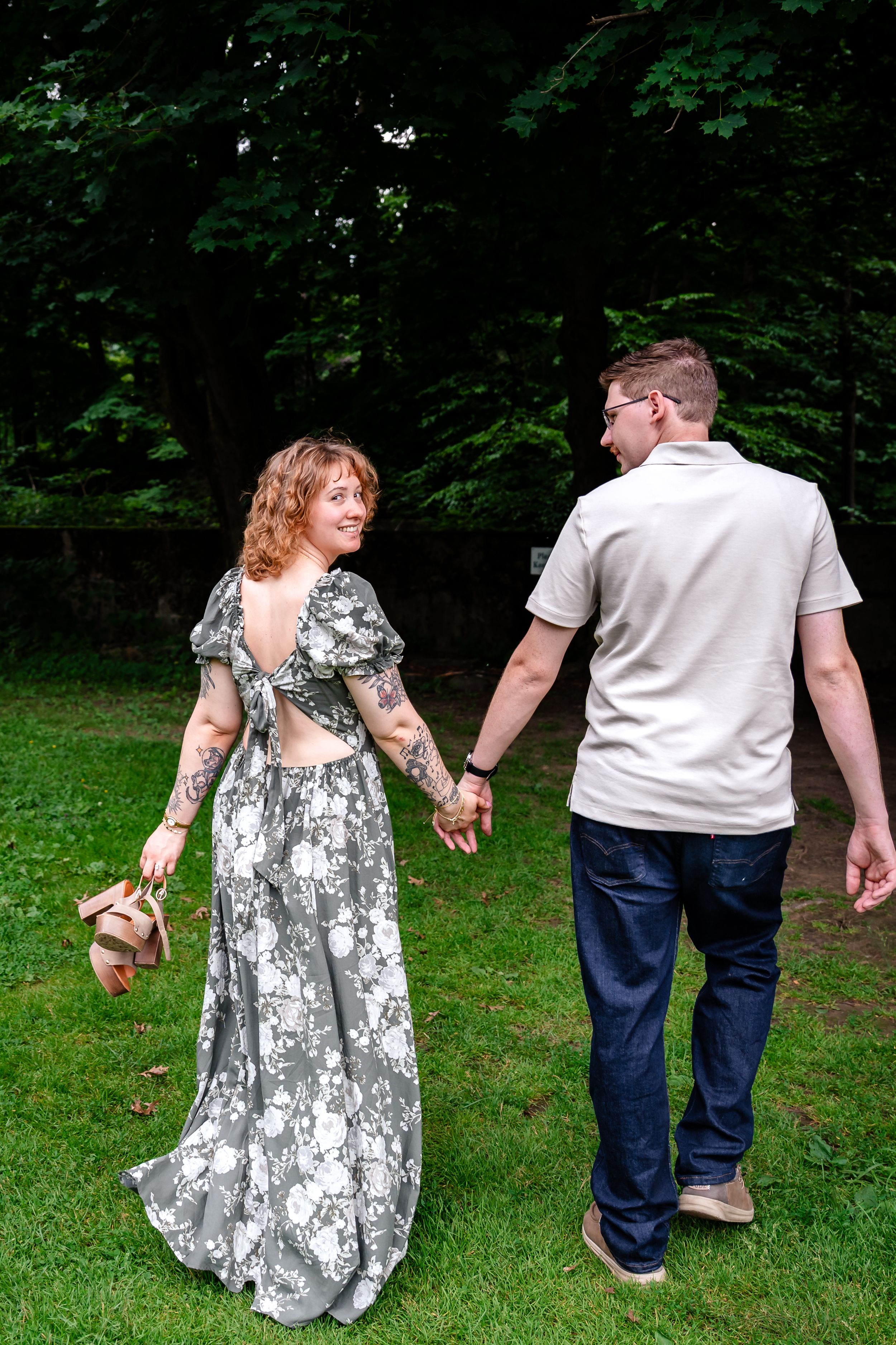 A couple walks hand-in-hand on grass, woman in floral backless dress with tattoos, man in casual attire, near green trees.