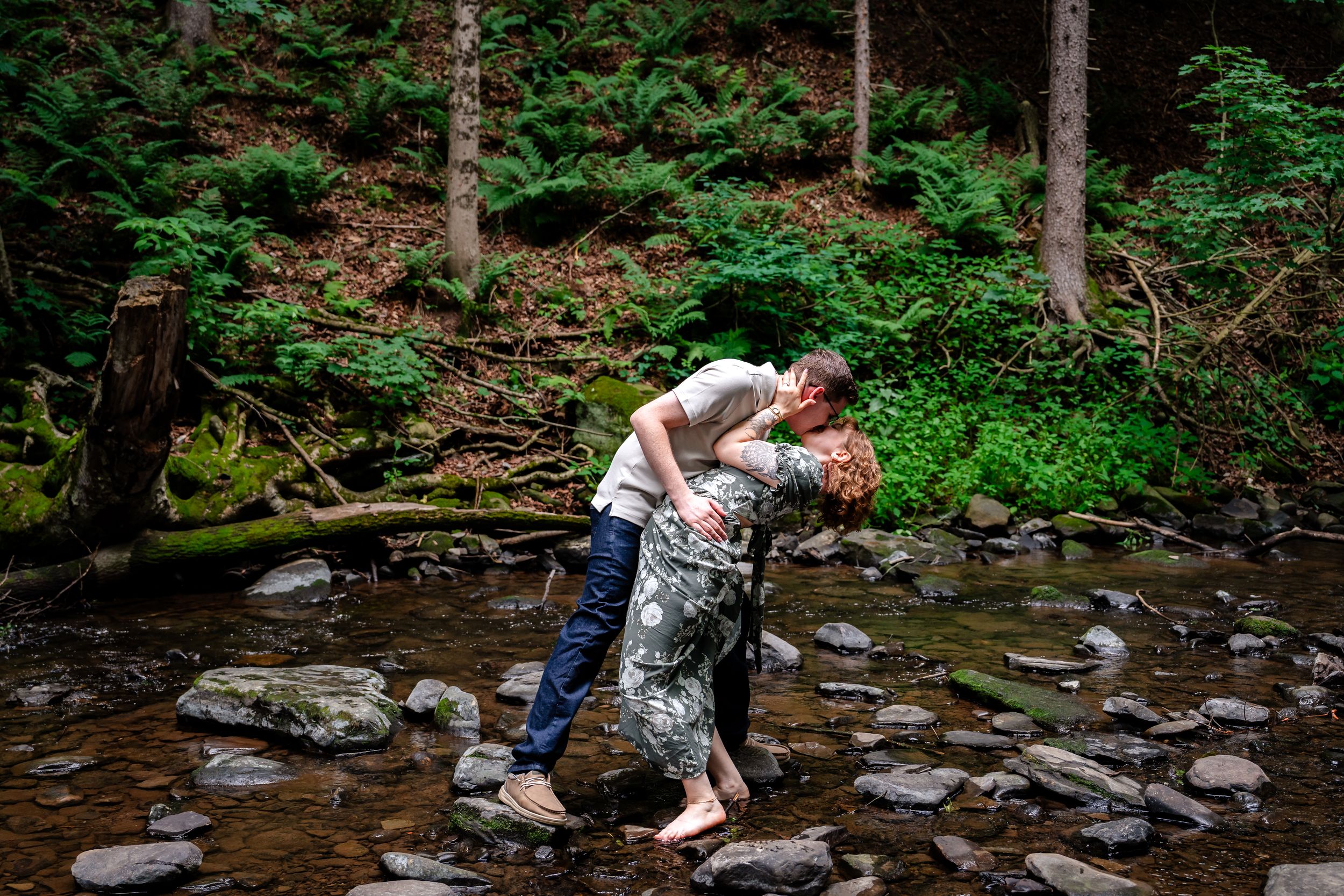 Couple sharing a romantic dip kiss while standing on rocks in a forest creek surrounded by lush green ferns.