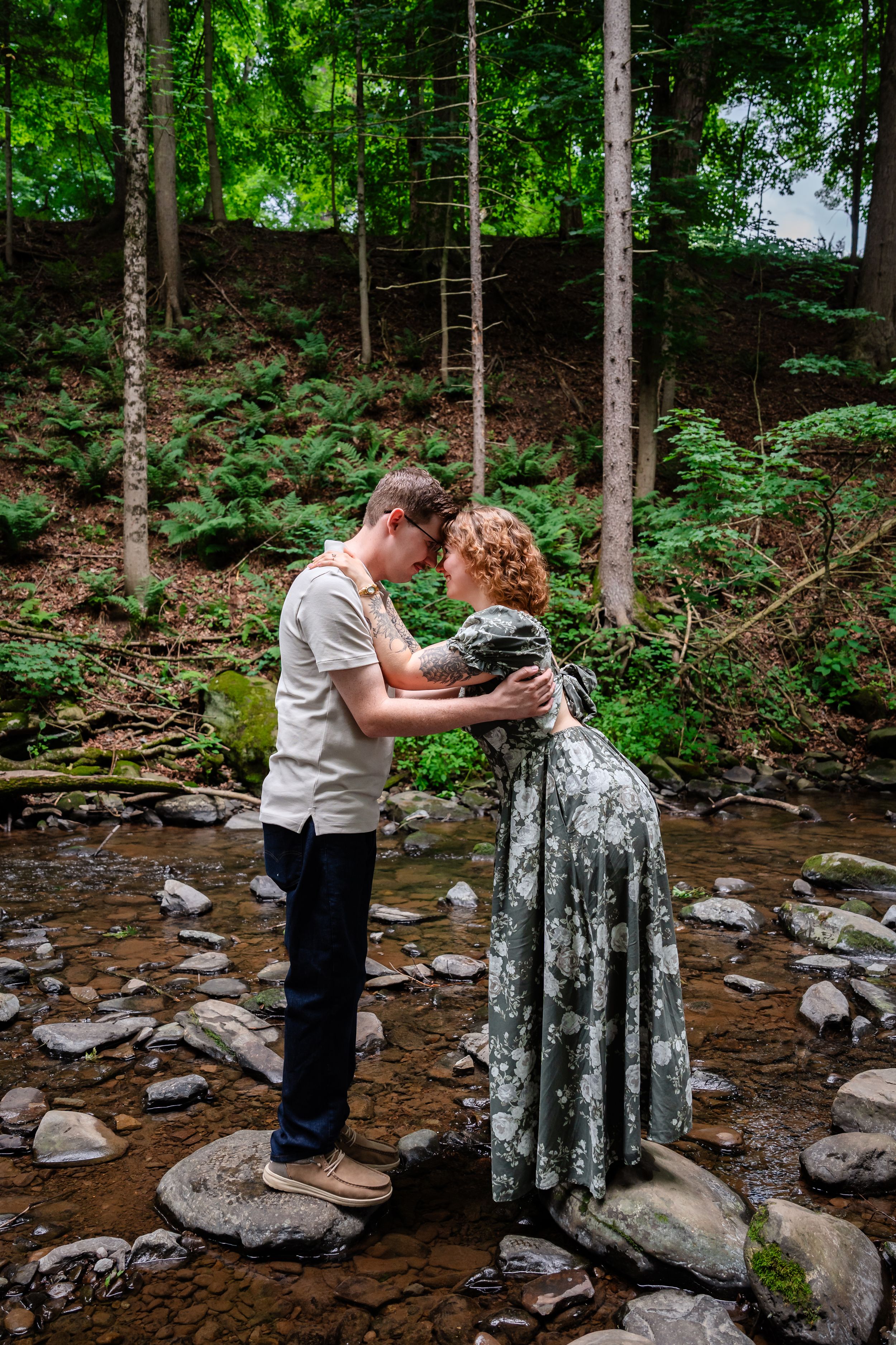 Couple sharing a romantic moment on rocks by a forest stream, woman in floral dress, lush green woodland backdrop.
