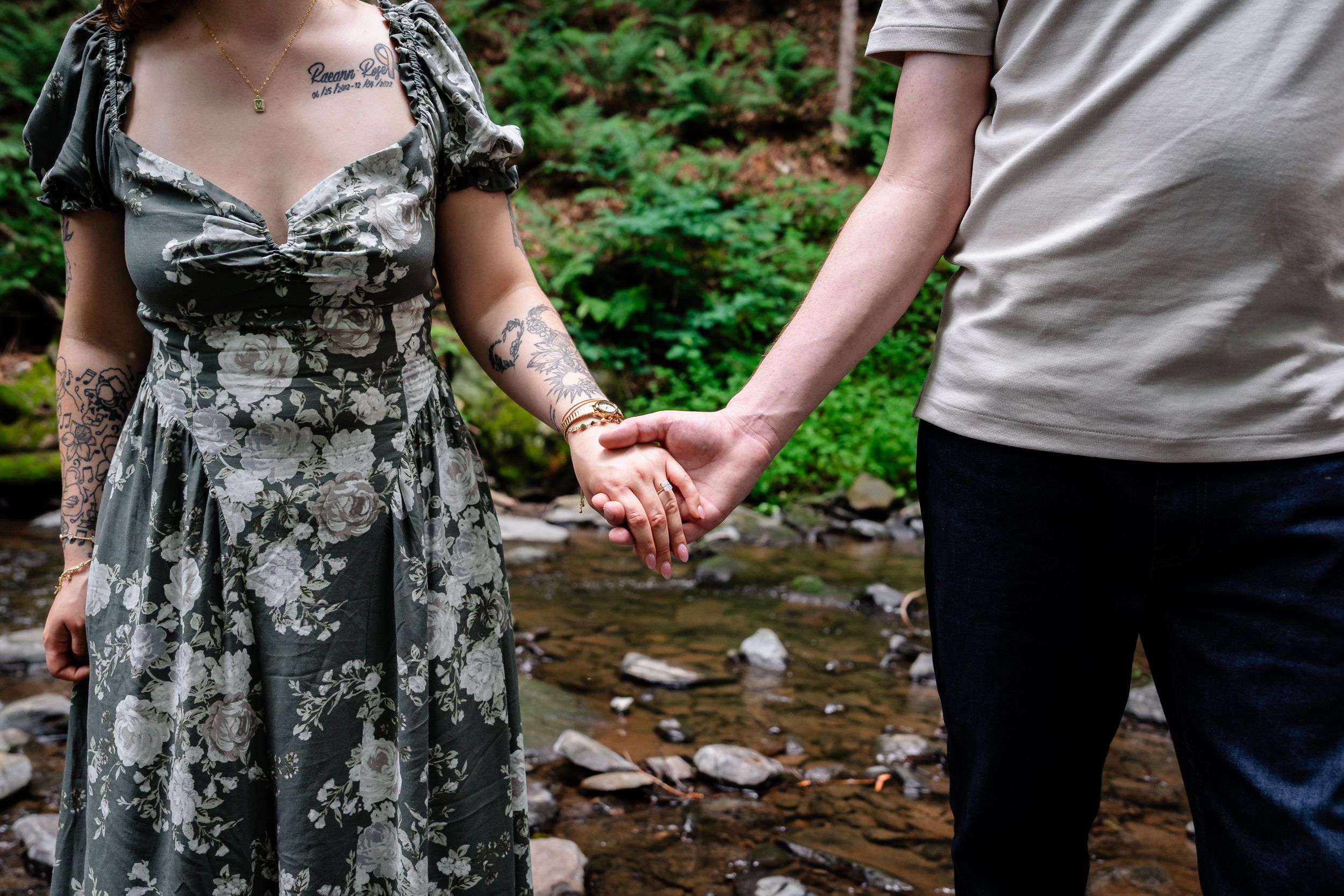 Couple holding hands near a rocky creek, woman in floral dress with tattoos, man in gray shirt and dark pants.
