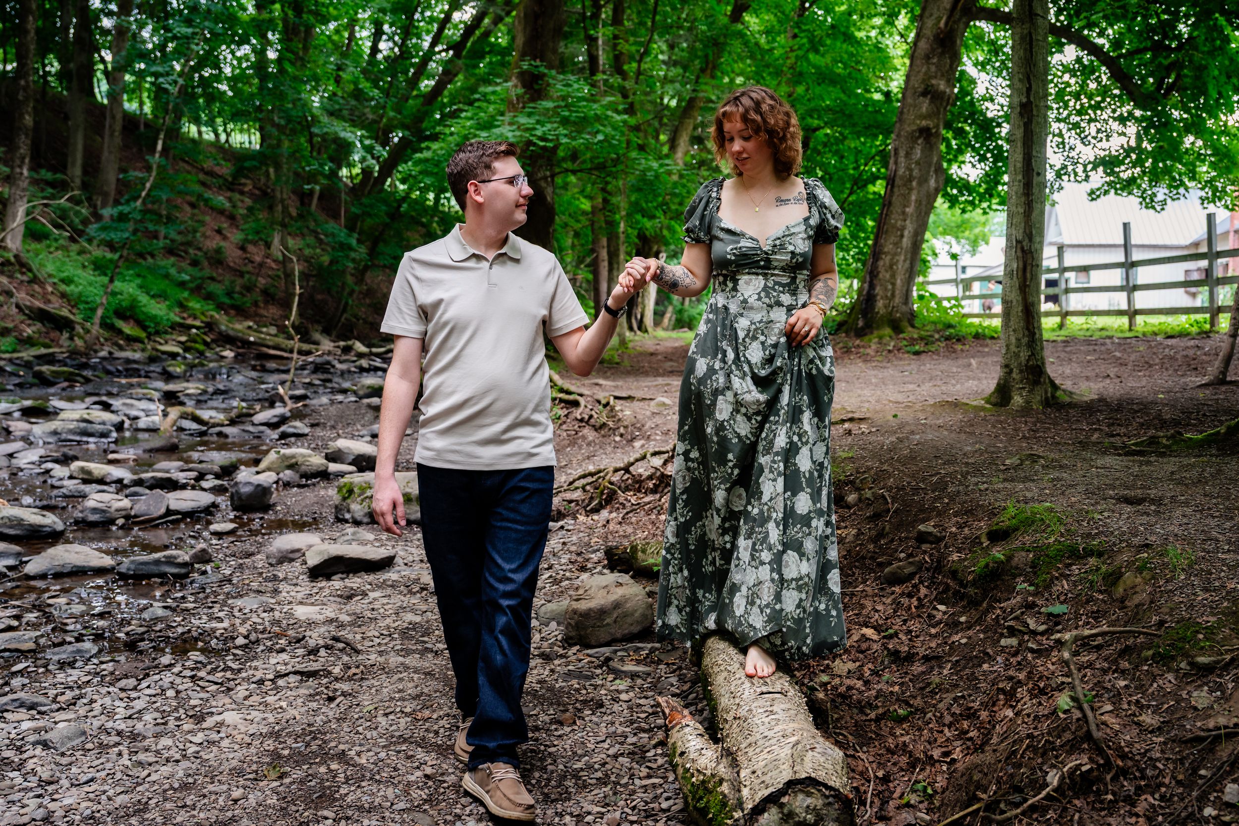 A couple walks hand in hand through a lush green woodland path, smiling at each other during an outdoor photoshoot.
