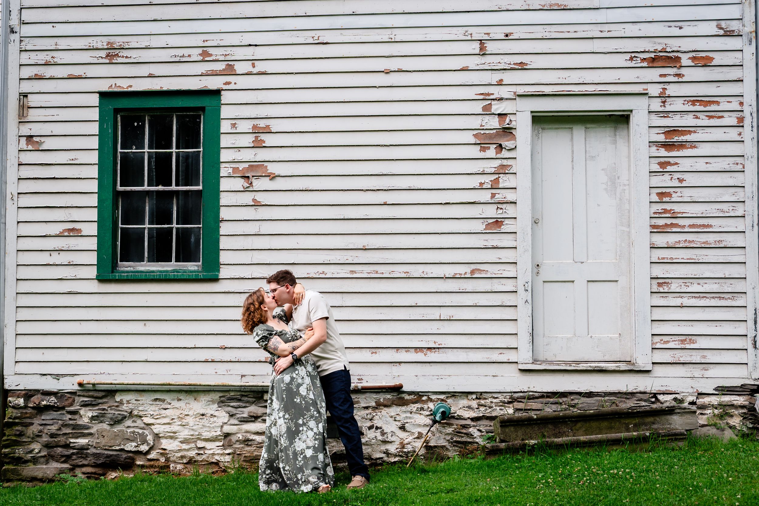 Couple sharing a romantic kiss embrace in front of a rustic white wooden building with peeling paint and green trim.