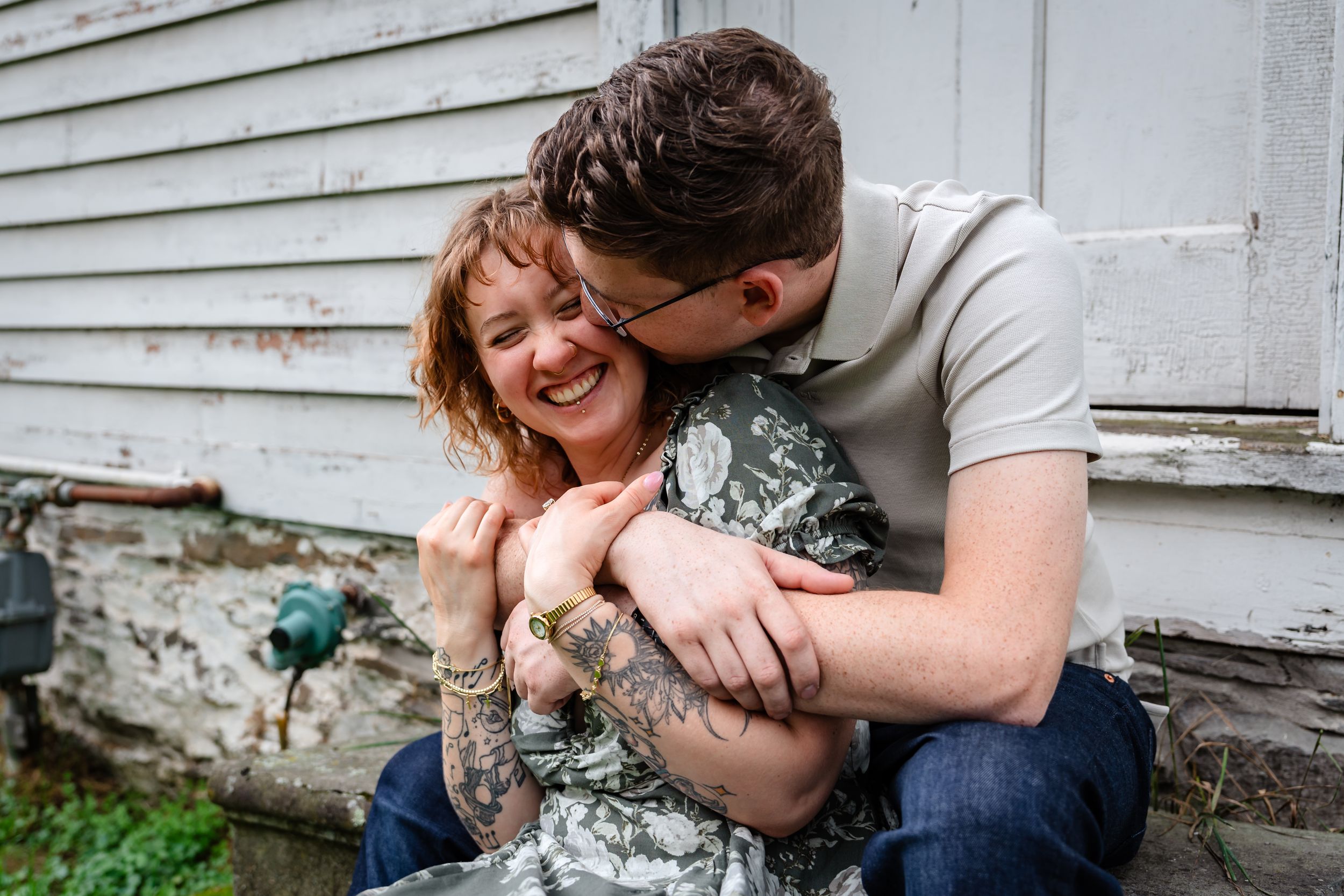 A couple shares a tender moment, man kissing woman's cheek as she laughs joyfully on rustic steps.