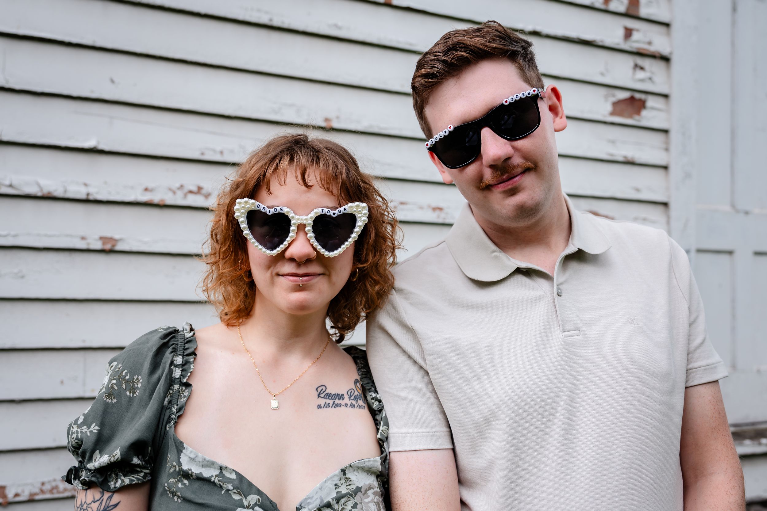 A couple posing together, woman wearing decorative heart-shaped sunglasses and floral dress, man in beige polo with dark sunglasses.