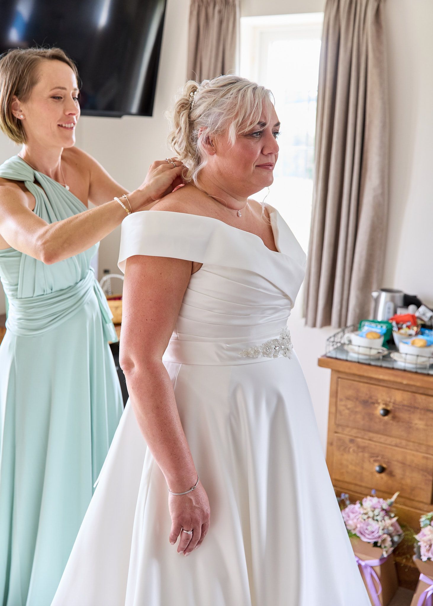 Bridesmaid in mint dress helping bride in white off-shoulder gown with necklace clasp in bright hotel room at white hart inn Derbyshire