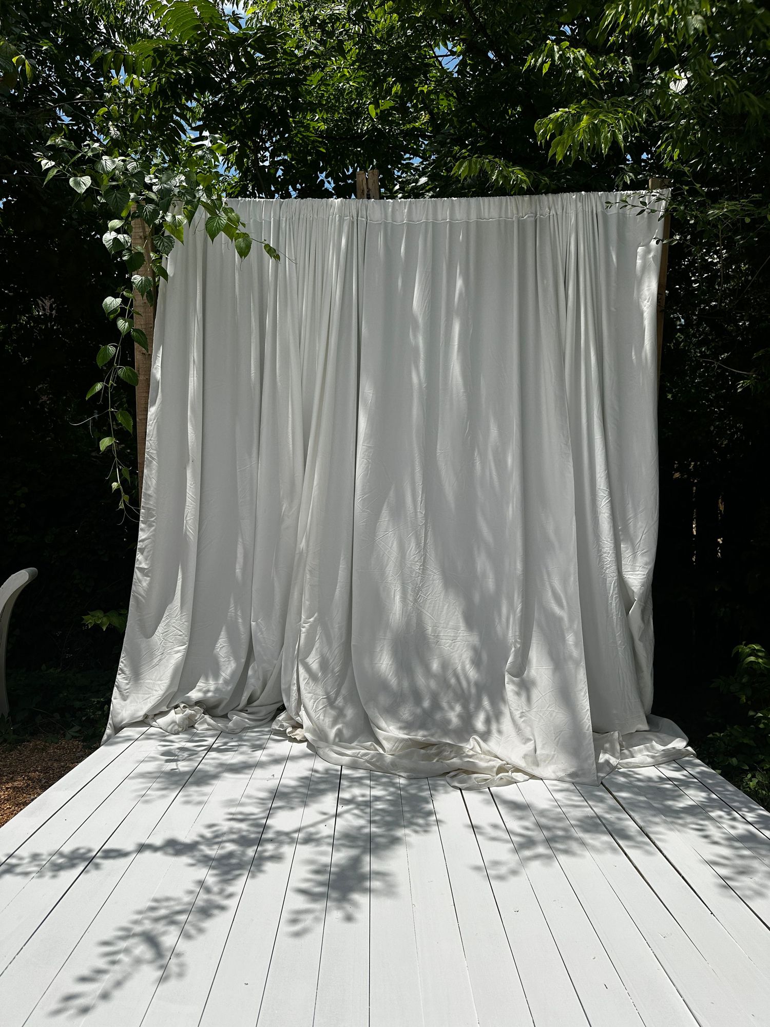White draped fabric backdrop on outdoor wooden deck surrounded by lush green trees with dappled sunlight.