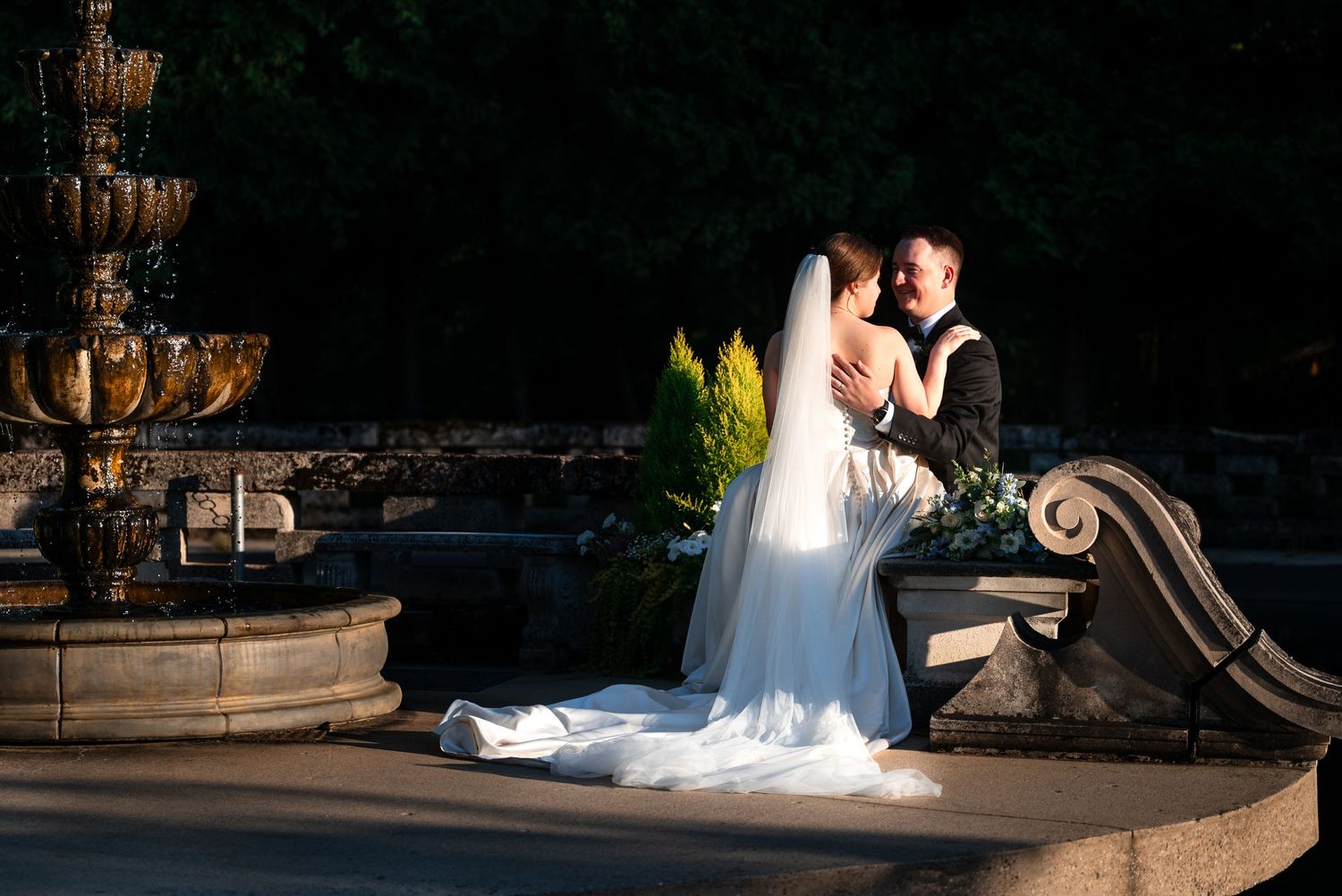 Bride and groom share a romantic embrace near a stone fountain at golden hour in an outdoor wedding setting.