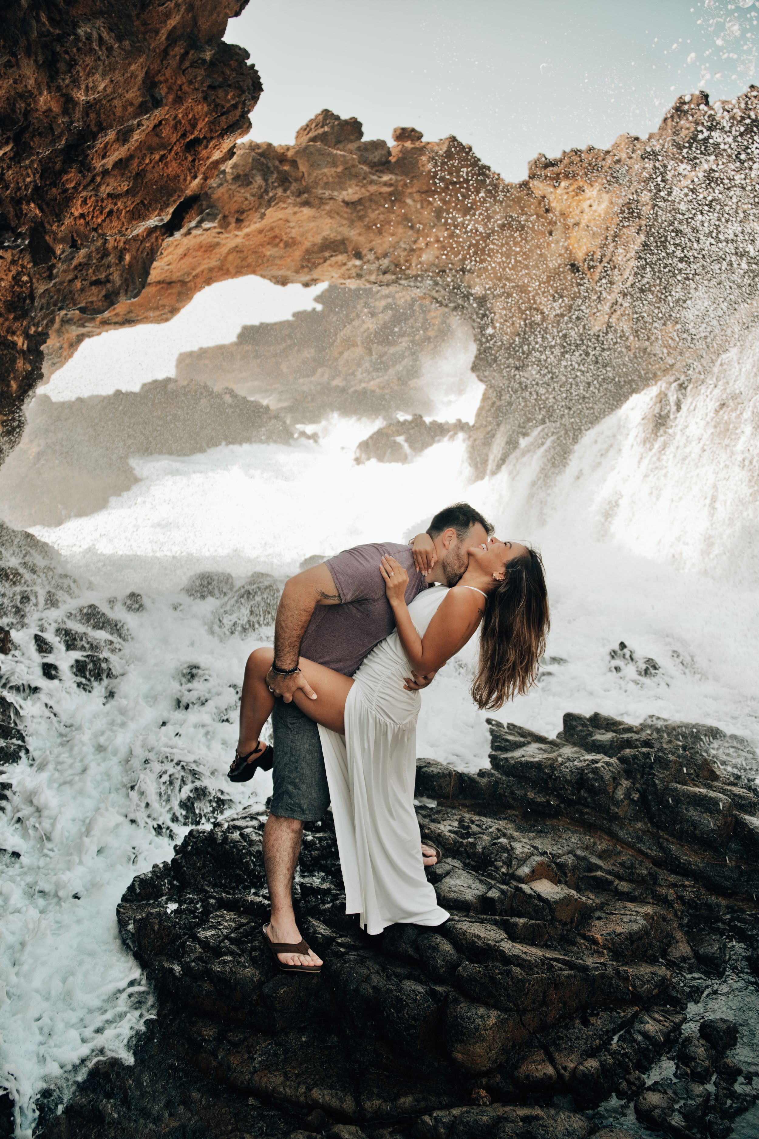 Couple kissing on rocky shore with ocean waves crashing through natural rock arch behind them.