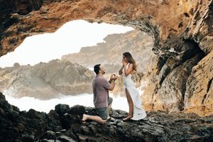 Man proposes to woman on rocky coastal cave with ocean waves in background during romantic outdoor engagement.