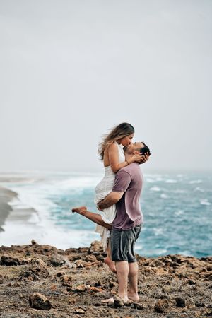 A romantic couple embraces on rocky coastal cliffs, man lifting woman as ocean waves crash behind them.