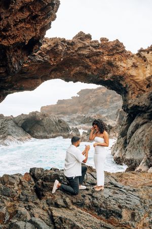 Man proposes to woman on rocky coastal arch with ocean waves, both dressed in white for a romantic seaside engagement.