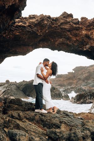 Couple kissing under a natural rock arch by the ocean, man in white shirt and woman in white dress.