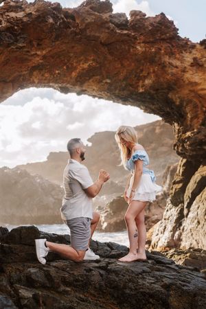Man proposes on one knee to woman under rocky arch by the ocean.
