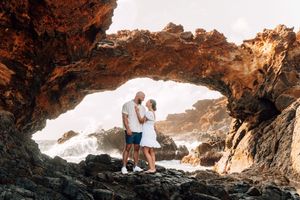 Couple standing under a natural rock arch by the ocean waves at sunset in a tropical coastal location.