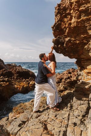 Couple kissing on rocky coastline with ocean waves and blue sky in the background.