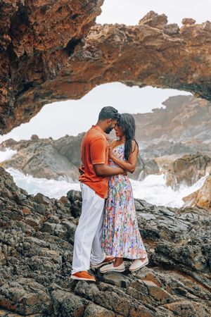 Couple embracing romantically on rocky coastal cliffs framed by natural rock arch with ocean waves behind them.