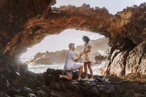 Man proposing to woman in a scenic rocky sea cave arch on a sunny beach, romantic ocean backdrop.
