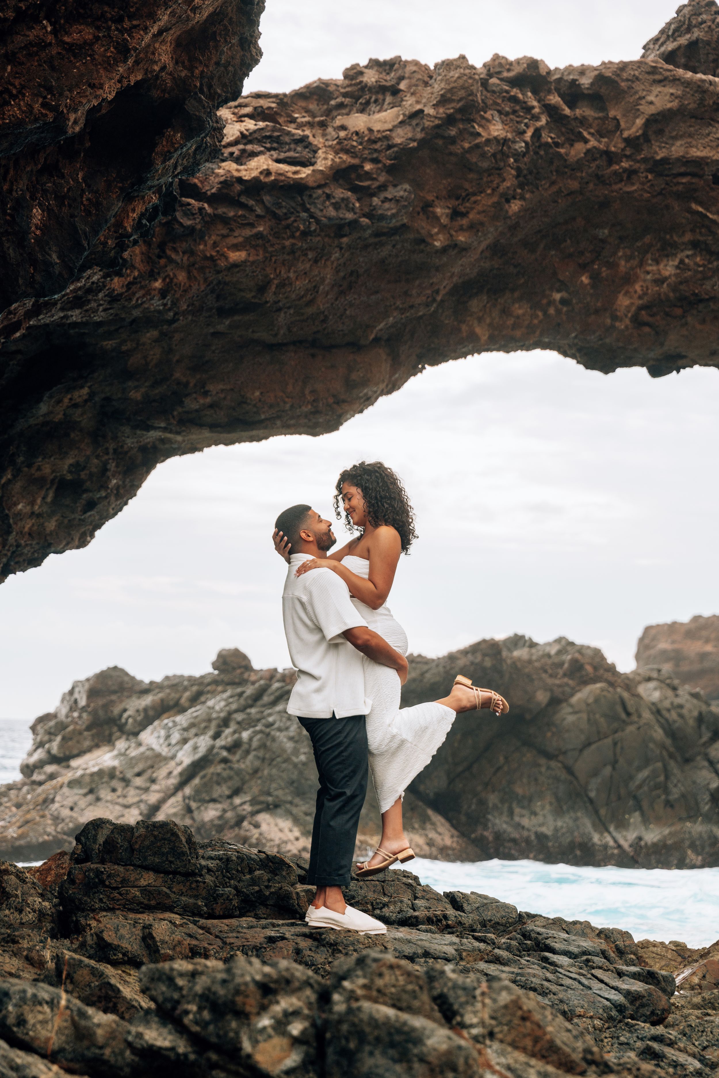 Couple embracing romantically under a natural rock arch on a rocky coastline with ocean waves in the background.